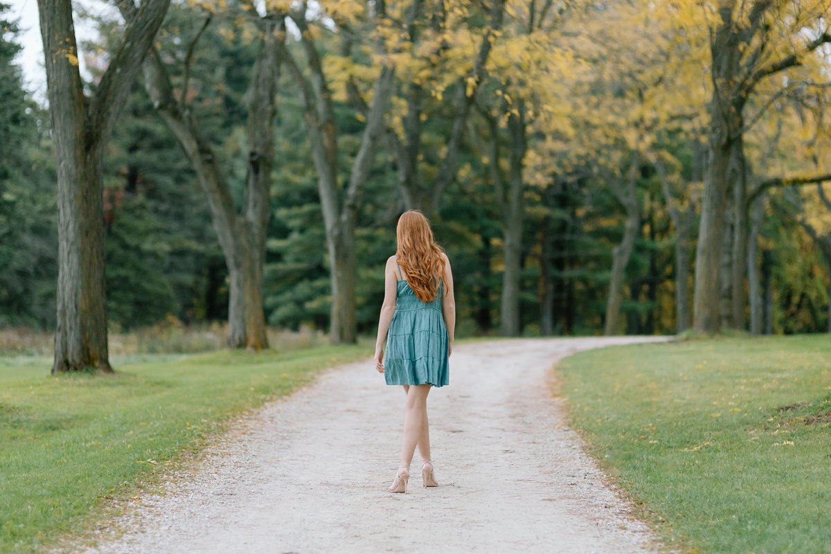 A girl with red hair and a teal blue dress walks along a pathway lined with trees and fall foliage.