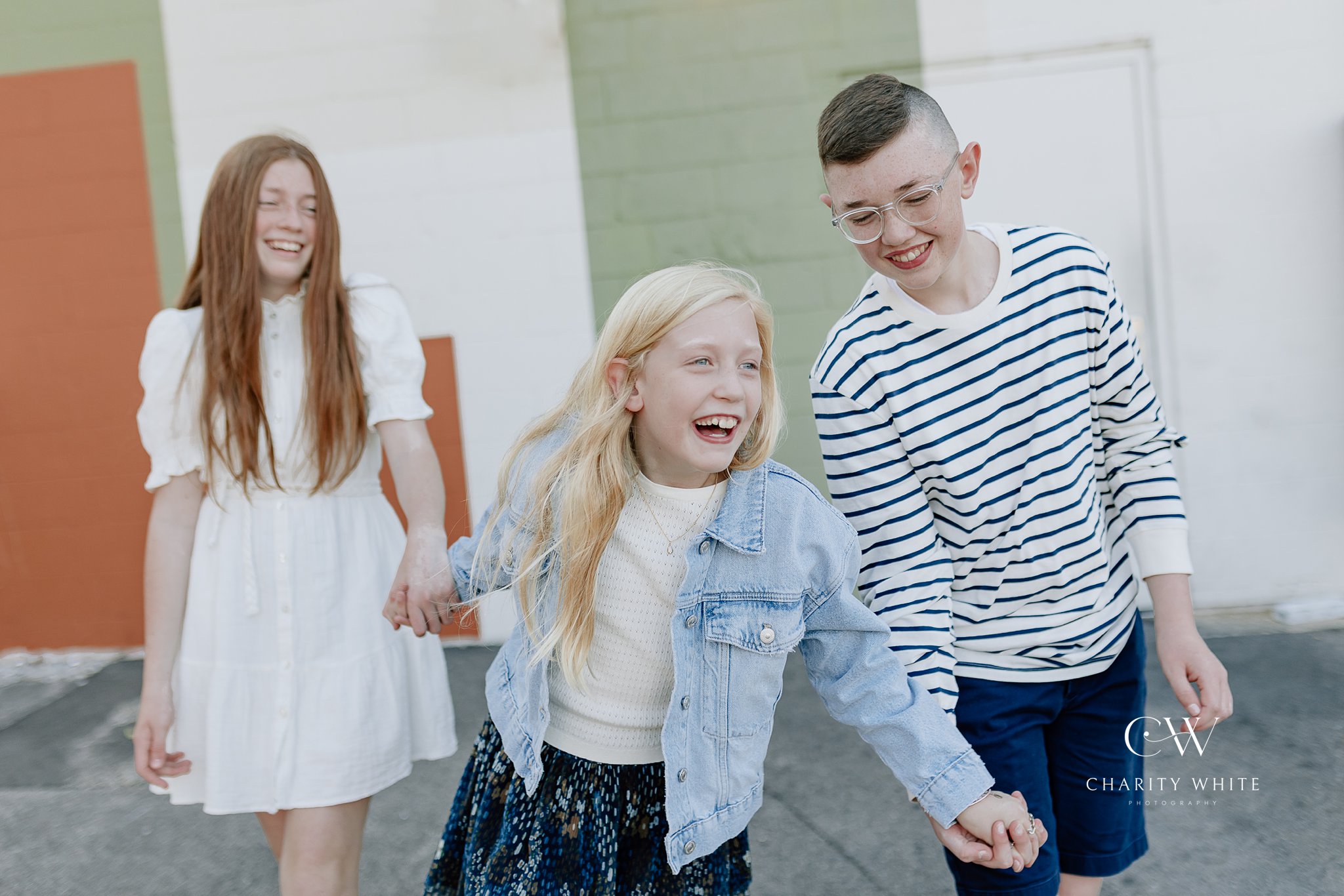 Two sisters and a brother hold hands and laugh together in a candid photo, wearing denim and navy blue shades against a painted alleyway wall in downtown Geneva, Illinois.