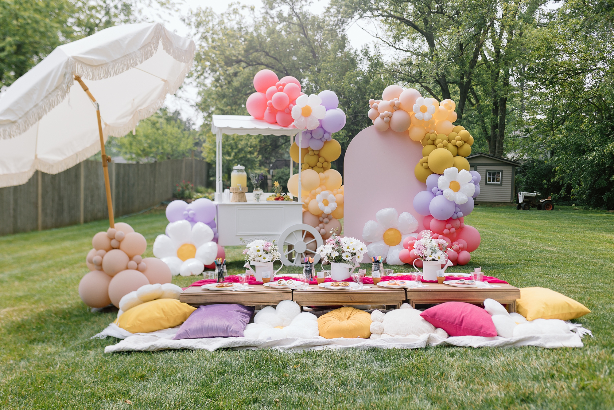 a daisy birthday party setup in backyard, with picnic spread, umbrella, white snack cart, and balloon arch with bright pink, yellow, lavender colors