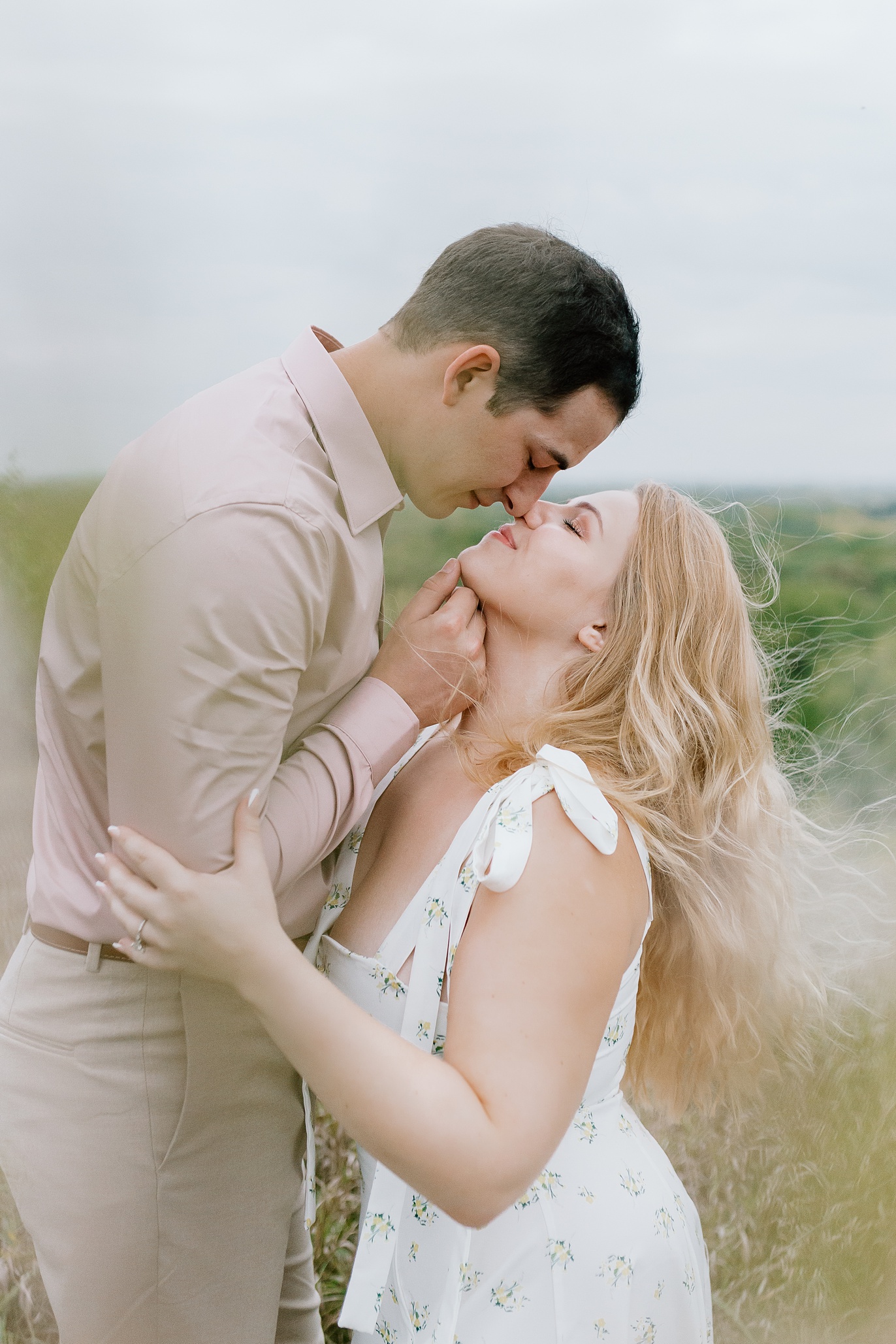 A young couple stand closely at the top of a hill and touch noses while wind blows in the girls hair. Engagement photos at Blackwell Forest Preserve in Warrenville Illinois